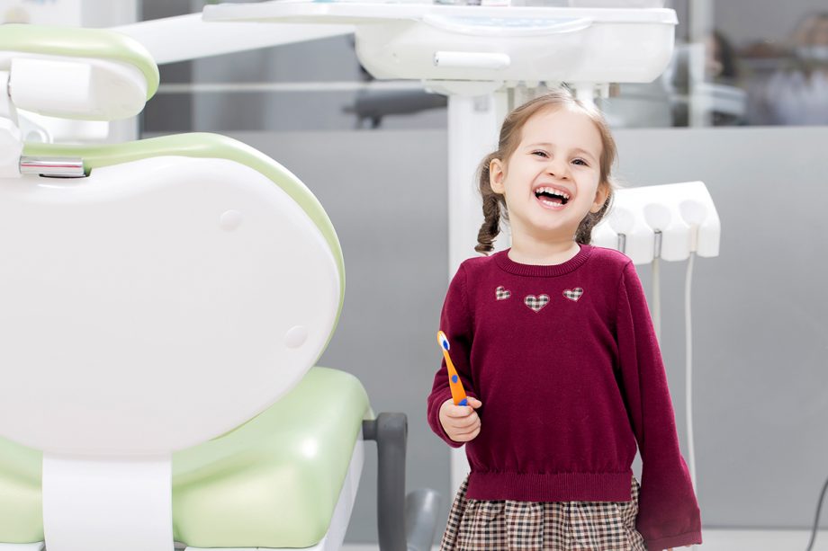 young girl next to dental chair smiling, holding toothbrush