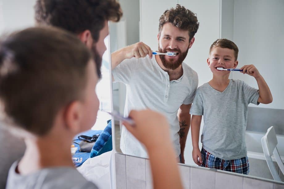 child and bearded man brush teeth in bathroom mirror